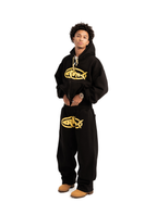 A person wearing a black hoodie with a gold logo on the front and black pants with a gold logo on the left leg, standing against a white background.