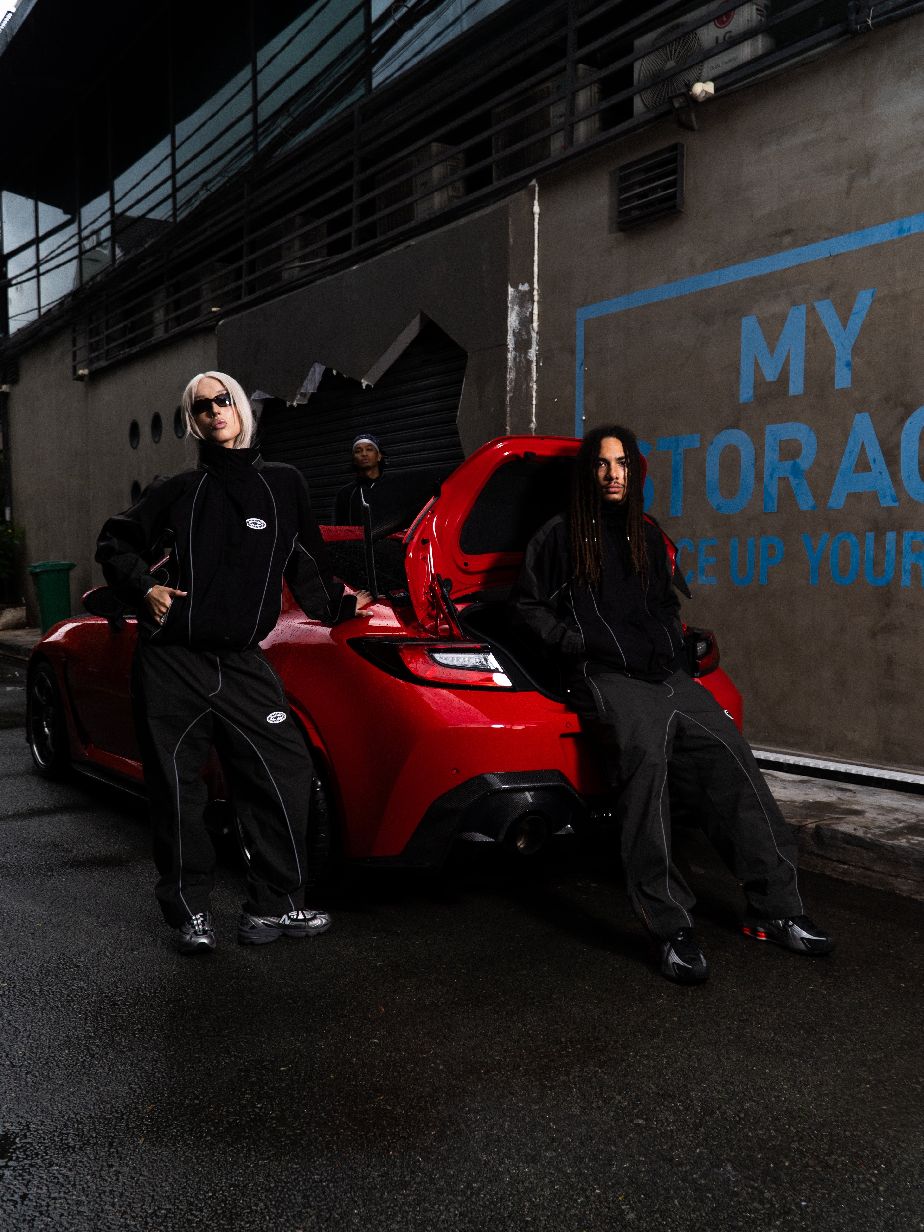 Two people sitting on a red sports car with a dark background and graffiti.