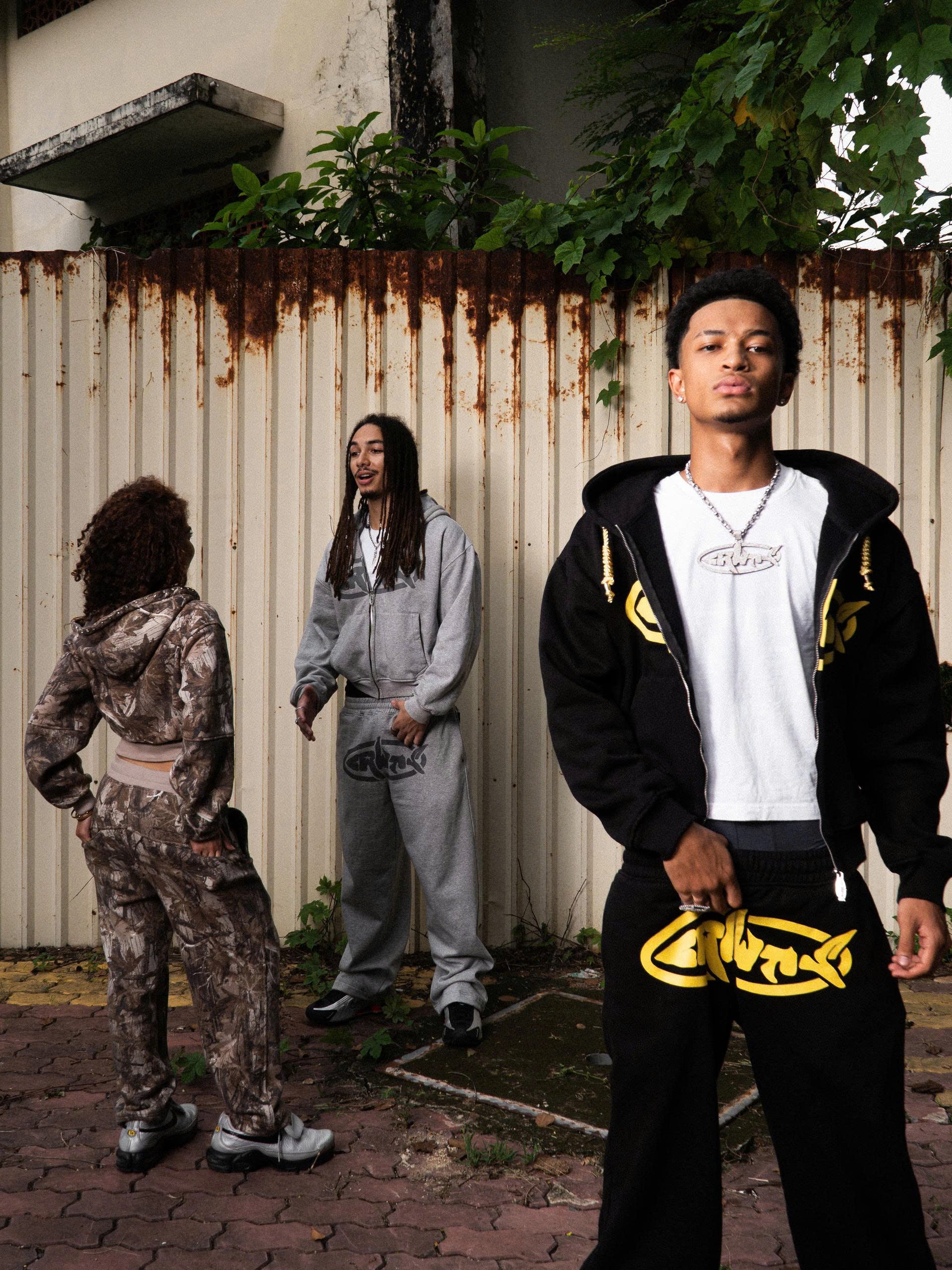 Three individuals standing outdoors with a rusty fence and plants in the background